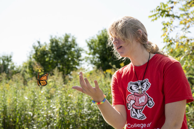 A student wearing a red Bucky Bader shirt holds out their hands as a monarch butterfly takes flight.