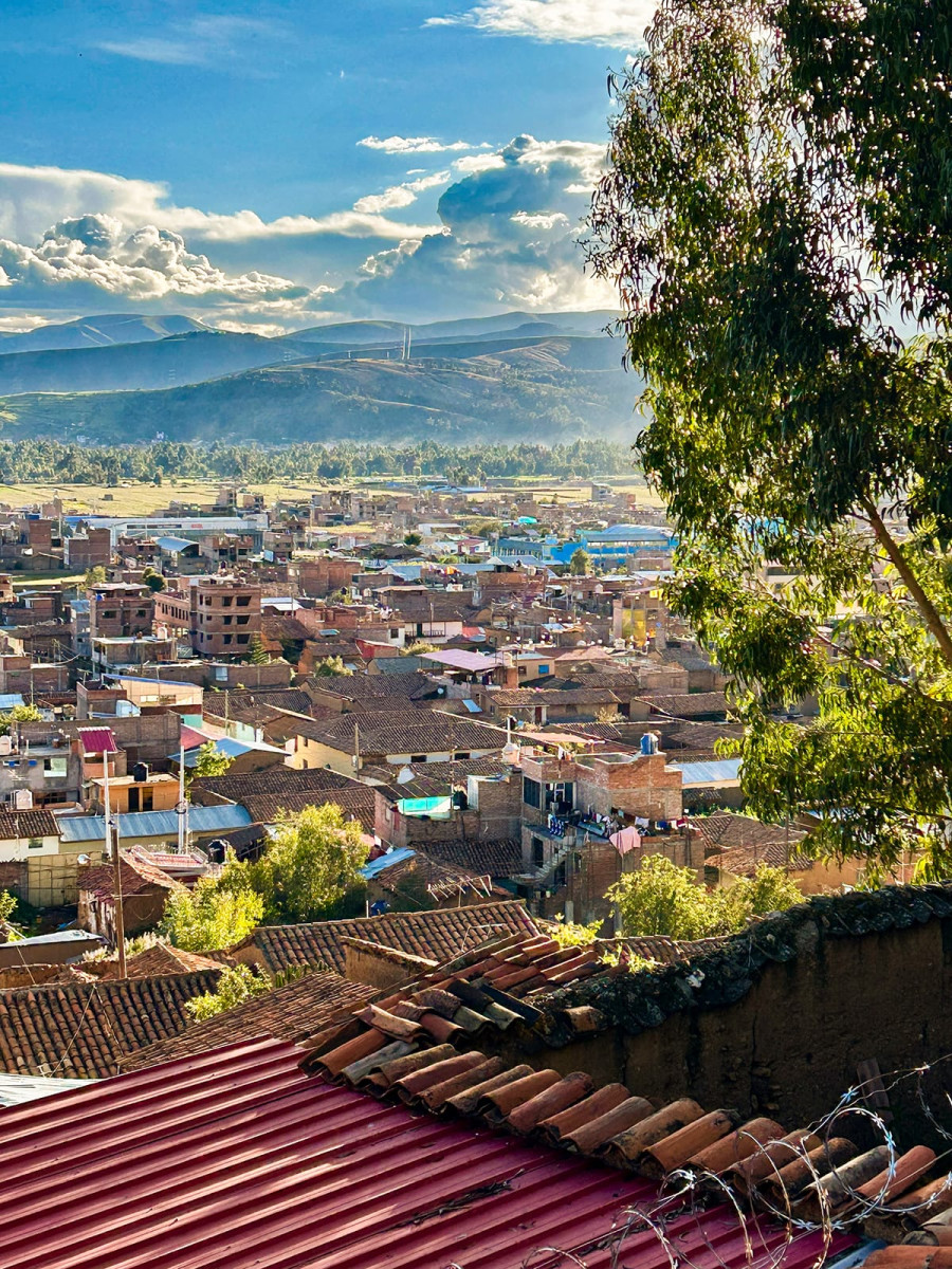 High-angle view overlooking a hillside town with terracotta-tiled roofs, surrounding green fields, and distant mountains beneath a partly cloudy blue sky.