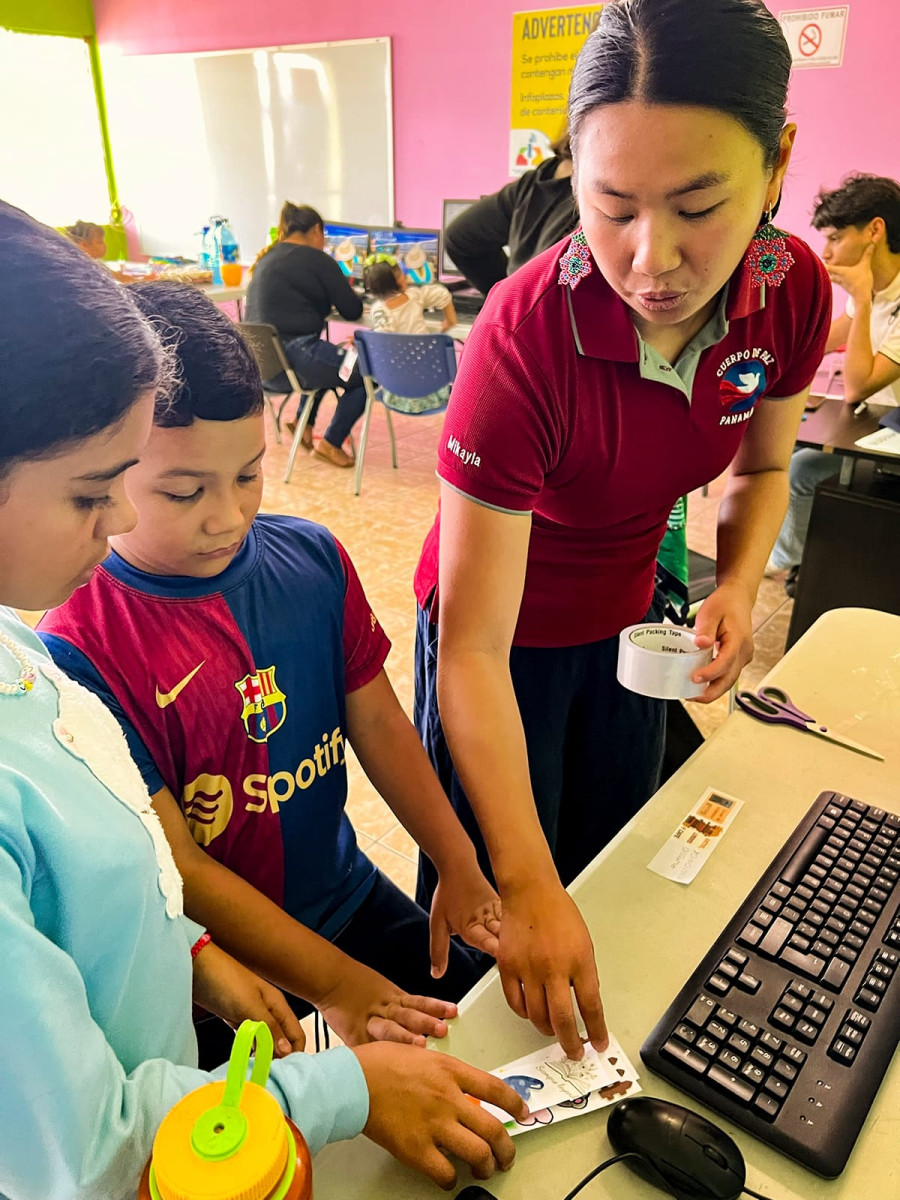 A Peace Corps volunteer wearing a red shirt holds a roll of tape while assisting two young students working at a computer desk.
