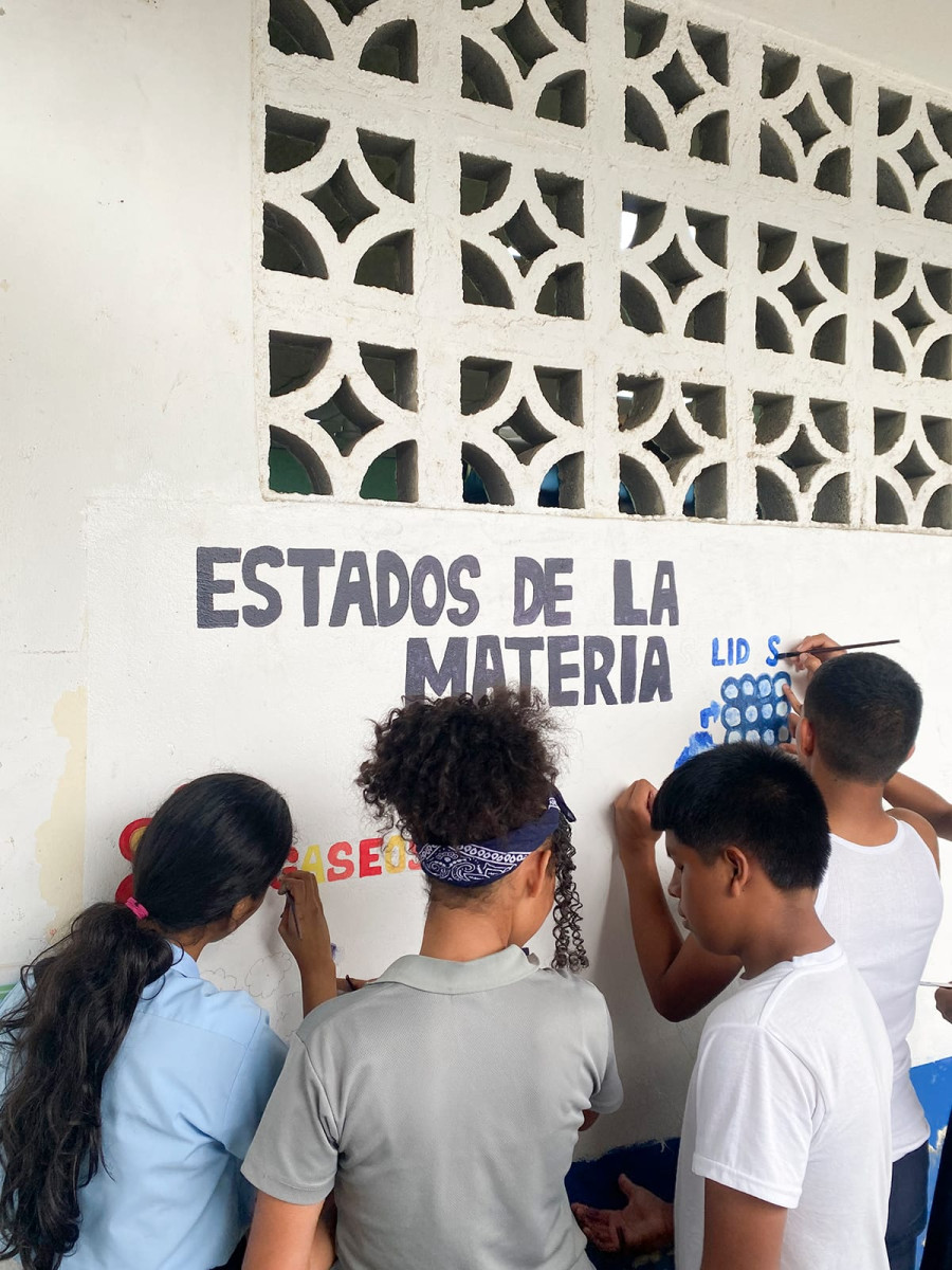 Four students stand facing a white wall while using paintbrushes to create an educational mural titled 'Estados de la Materia' ('States of Matter' in English).