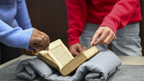 Two students page through an old book that rests on a cushion and table.