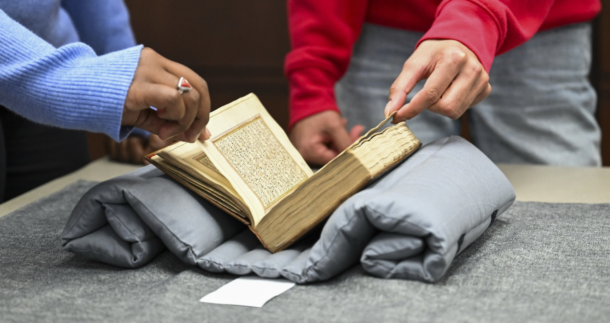 Two students page through an old book that rests on a cushion and table.