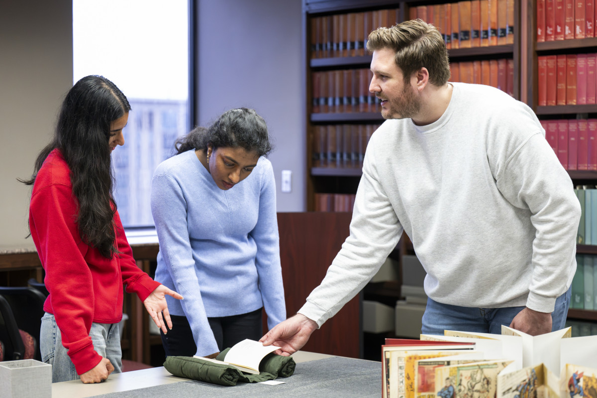 A graduate student assistant holds open a rare book and discusses it with two students.