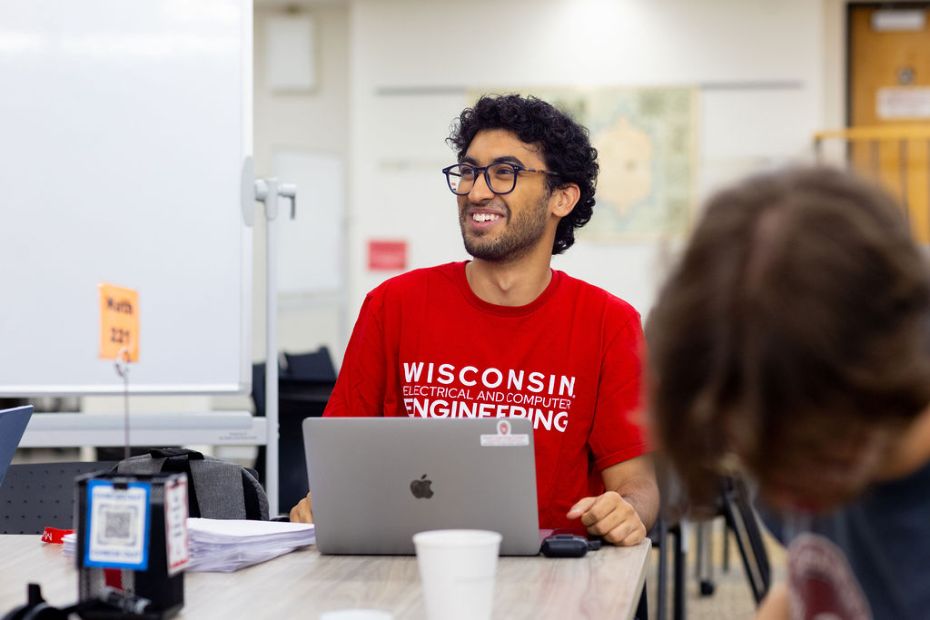 A man sits at a table with a laptop in front of him. He's wearing a Wisconsin Engineering tee shirt and is smiling to someone out of frame.