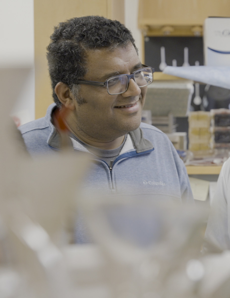A portrait of biochemistry professor Vatsan Raman in his lab, with equipment in the foreground.