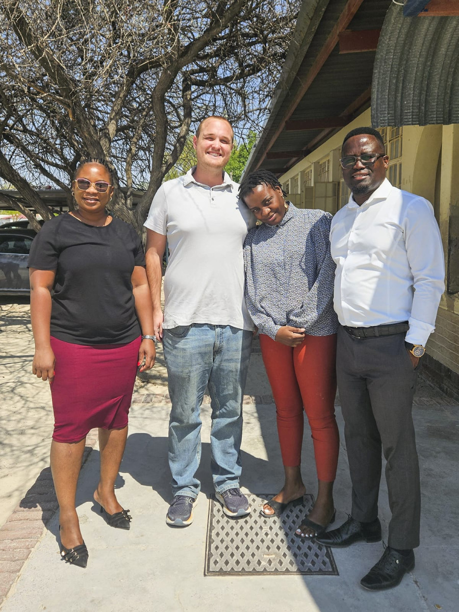 Four adults stand side-by-side outdoors near a building, smiling together for a group photo.
