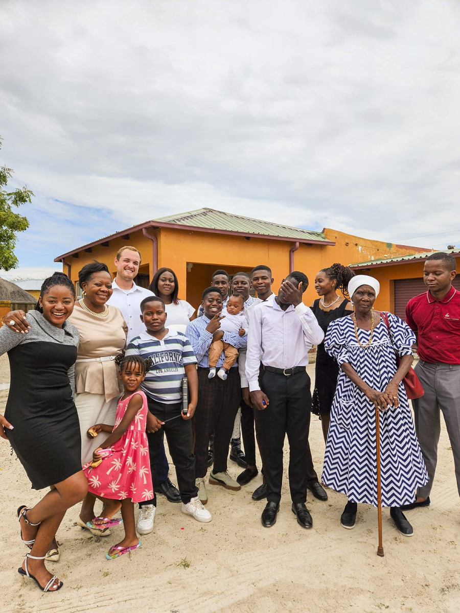 A large group of adults, children, and an elderly woman holding a cane pose together outside in front of a yellow-orange house.