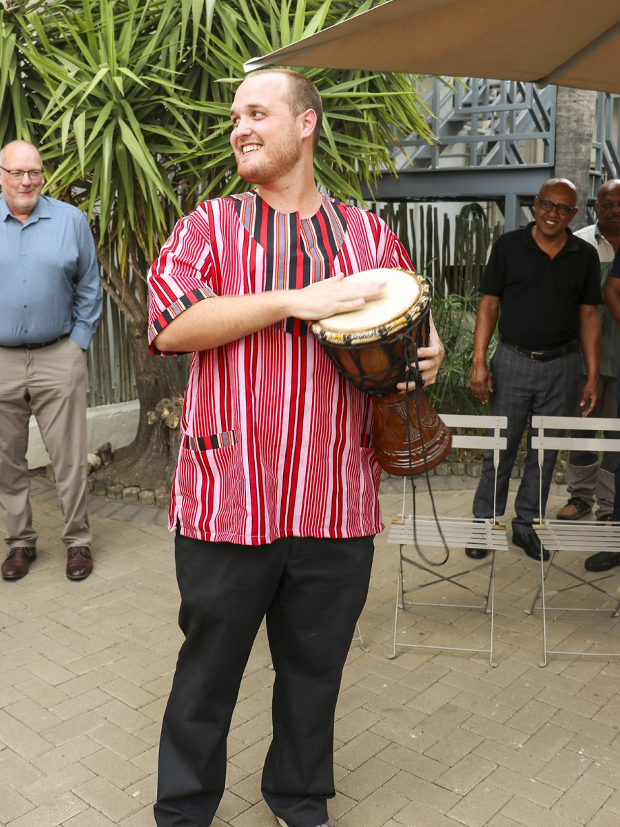 A smiling man wearing a red-and-black striped traditional tunic plays a wooden hand drum on an outdoor patio while onlookers watch.