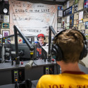 Johnny Raider wearing backwards baseball cap runs a live radio broadcast in the studio surrounded by microphones and radio equipment