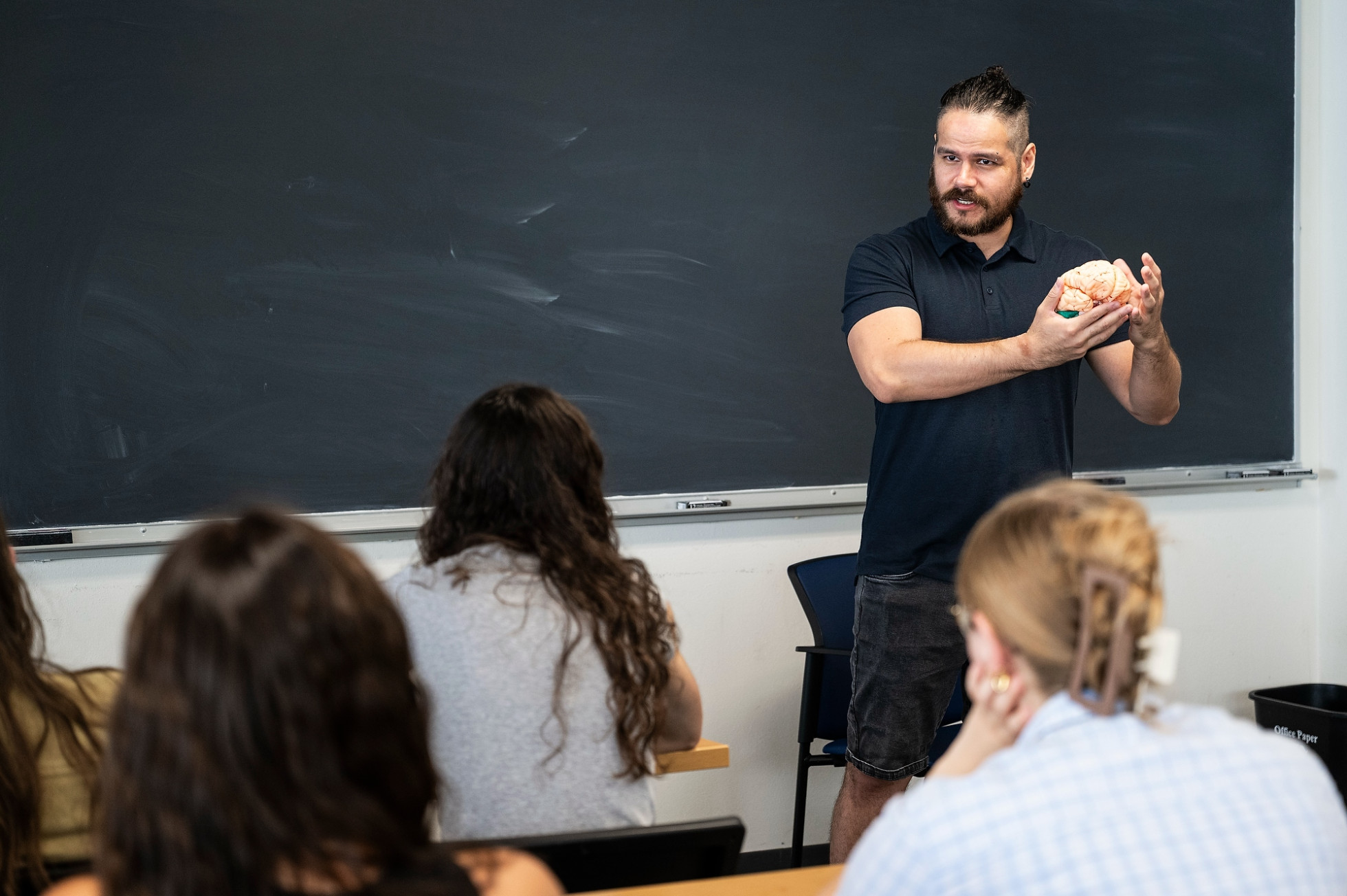 A professor in a black shirt talks with students in front of a blackboard.