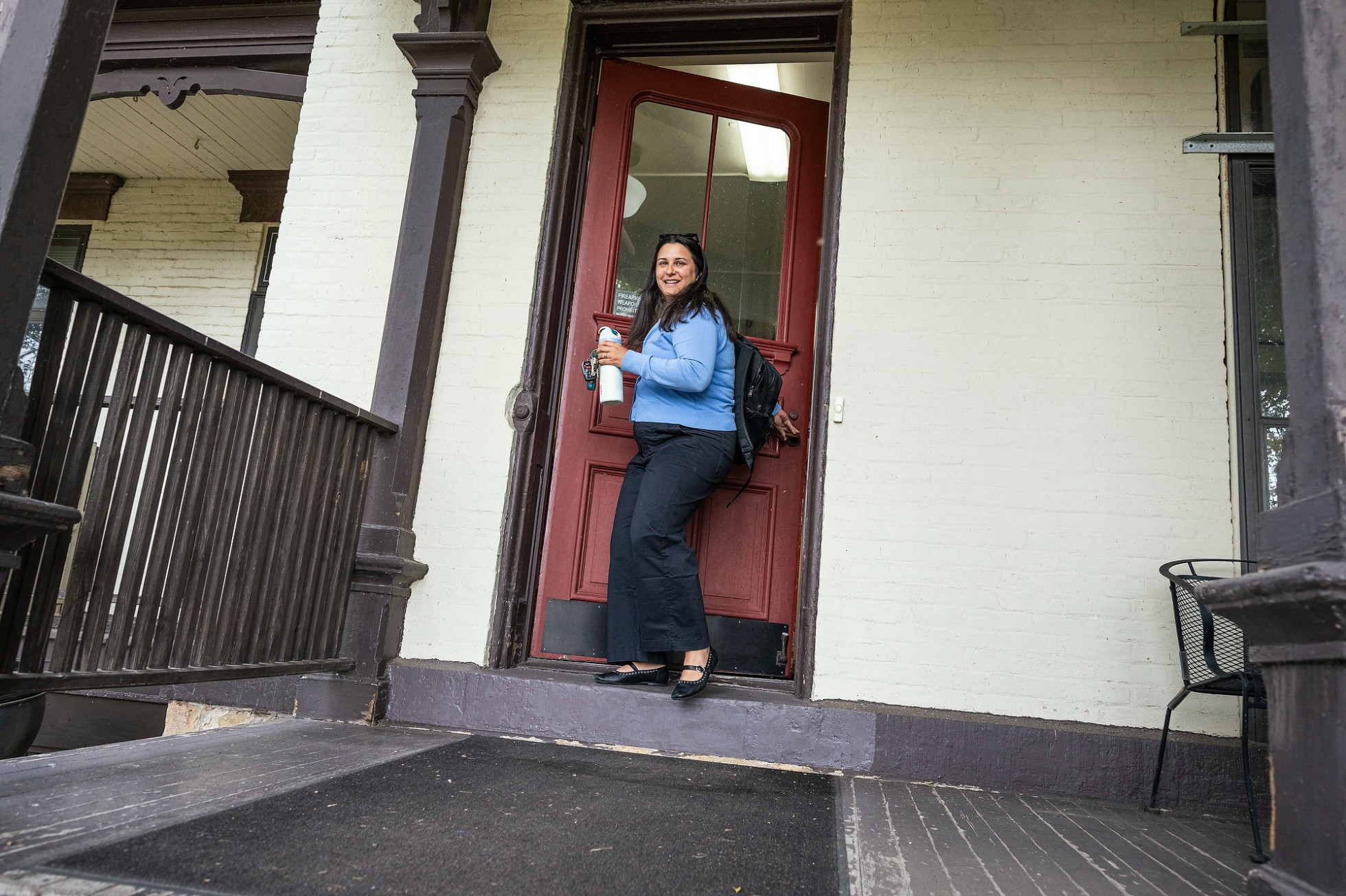 A person in a blue sweater holding a water bottle comes out of the exterior red door of a building.