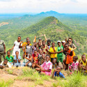 A large group of smiling children and adults pose together on a rocky hill summit overlooking an expanse of lush green countryside.