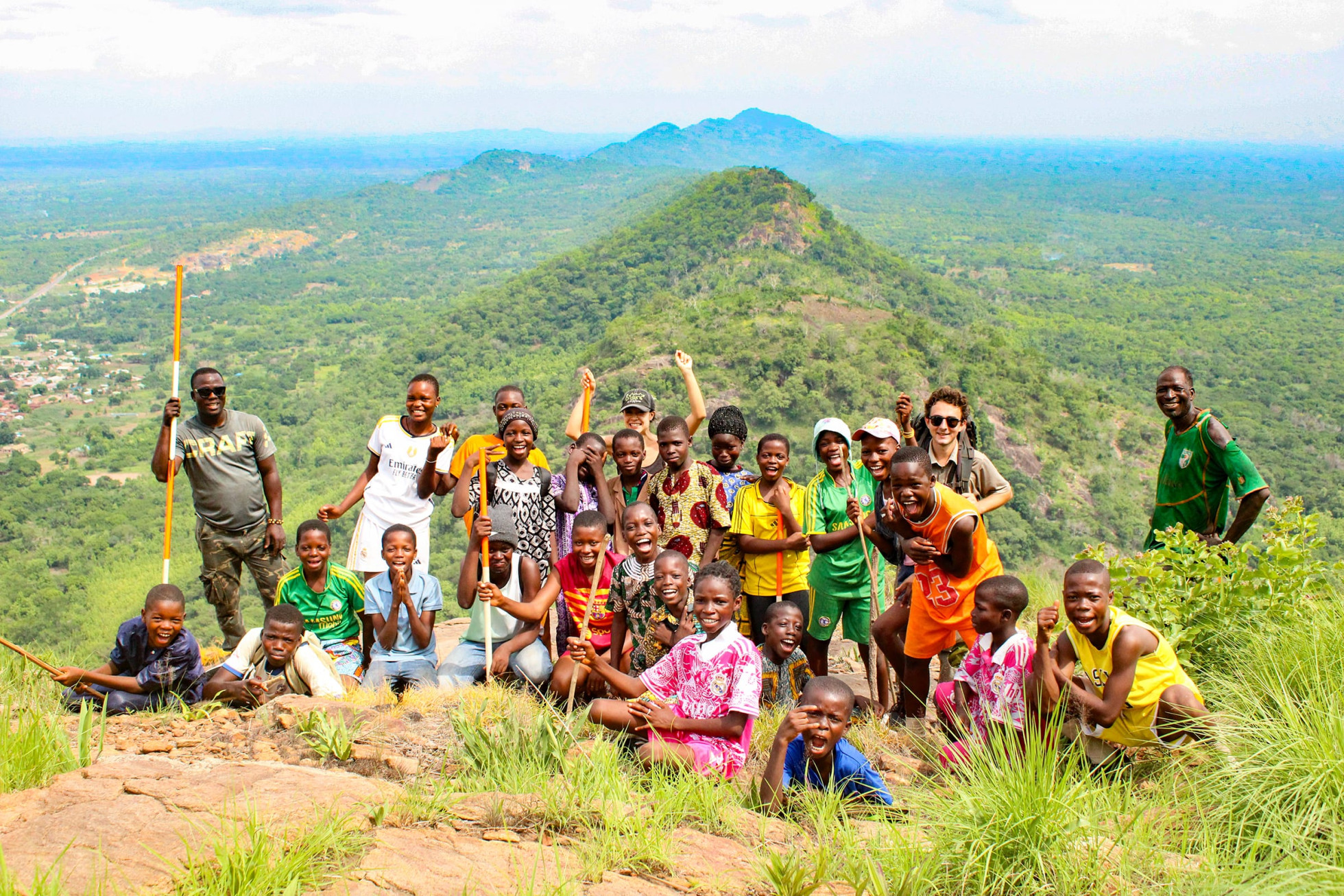 A large group of smiling children and adults pose together on a rocky hill summit overlooking an expanse of lush green countryside.