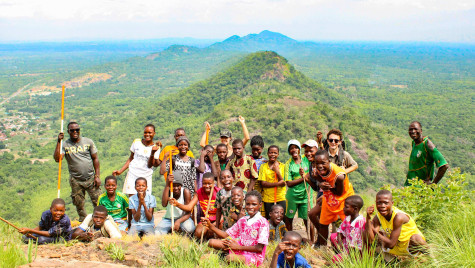 A large group of smiling children and adults pose together on a rocky hill summit overlooking an expanse of lush green countryside.