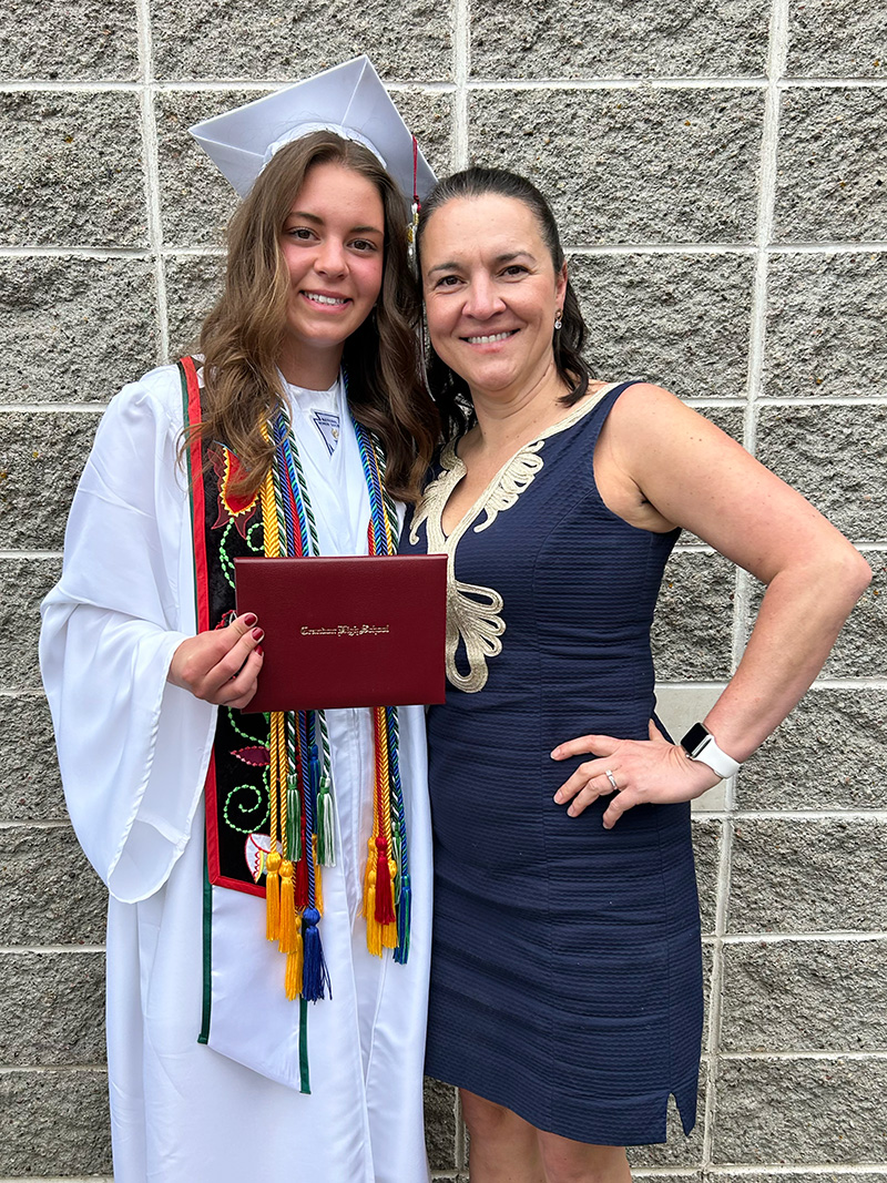 Student Maya Quade in graduation attire holding a diploma cover standing next to her mother