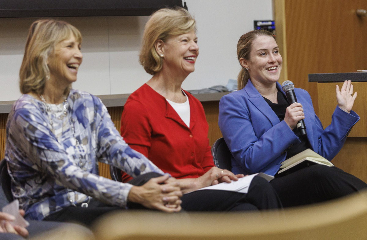 Senator Tammy Baldwin wears a red sweater and sits in between two women. One holds a microphone and addresses the group.