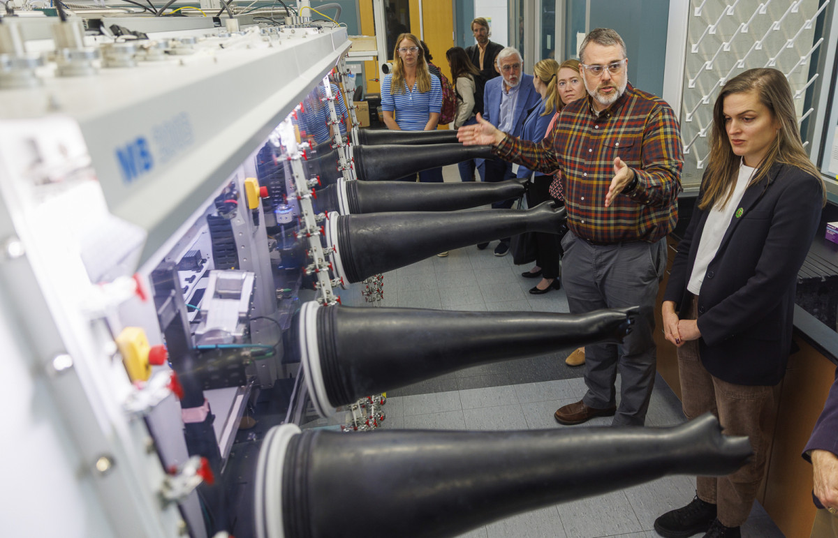 A group of people stand in front of a robotics machine while Botts explains its functionality to the group.