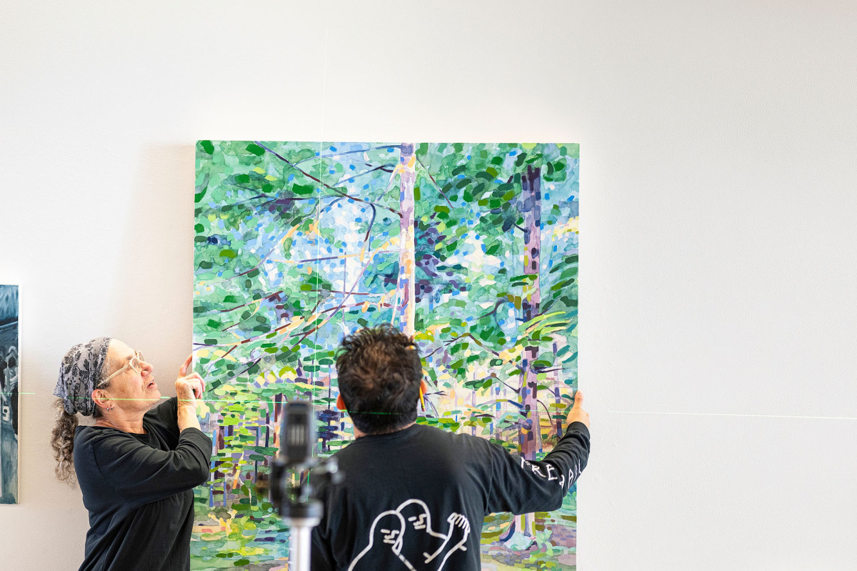 Laurie Beth Clark and Mark Almanza work to hang a painting of a green forest