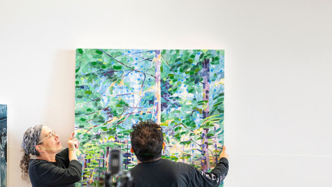 Laurie Beth Clark and Mark Almanza work to hang a painting of a green forest