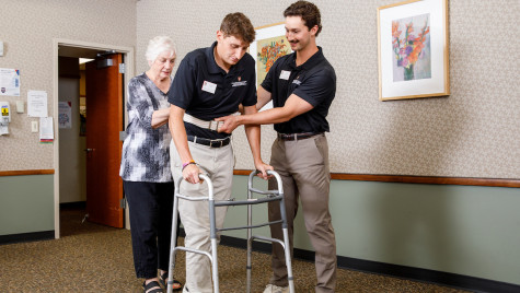 An older woman stands behind a young man wearing a gait belt and holding onto a walker, while another man stands beside them providing support in a clinic setting.