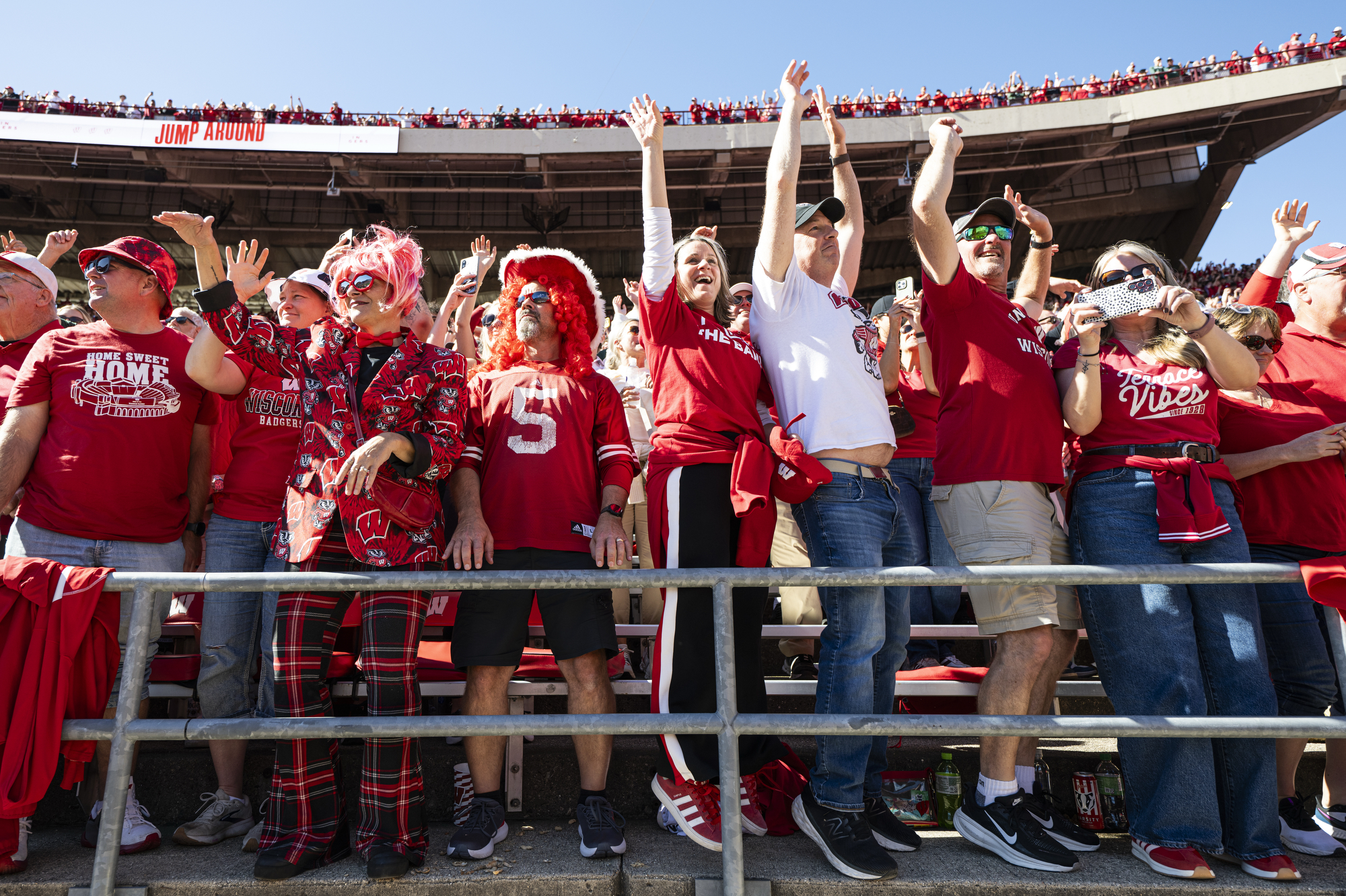 A group of Badger Fans in the stands at Camp Randall throw their hands in the air and jump around.