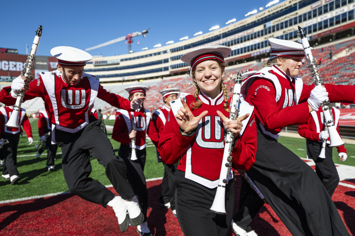 A member of the UW Marching Band holds her hands in a W formation as fellow band members jump in the air behind her.