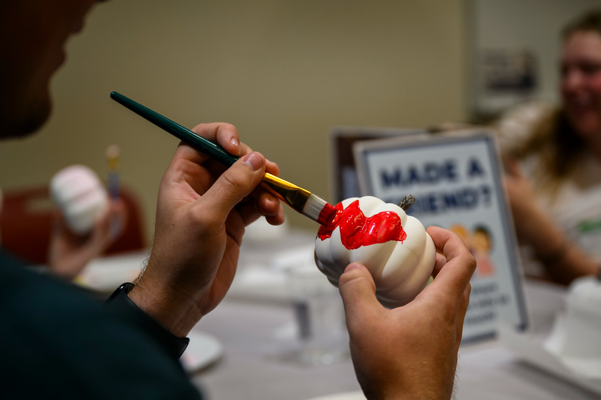 A close-up shows a student painting a red W on the side of a small, white pumpkin.