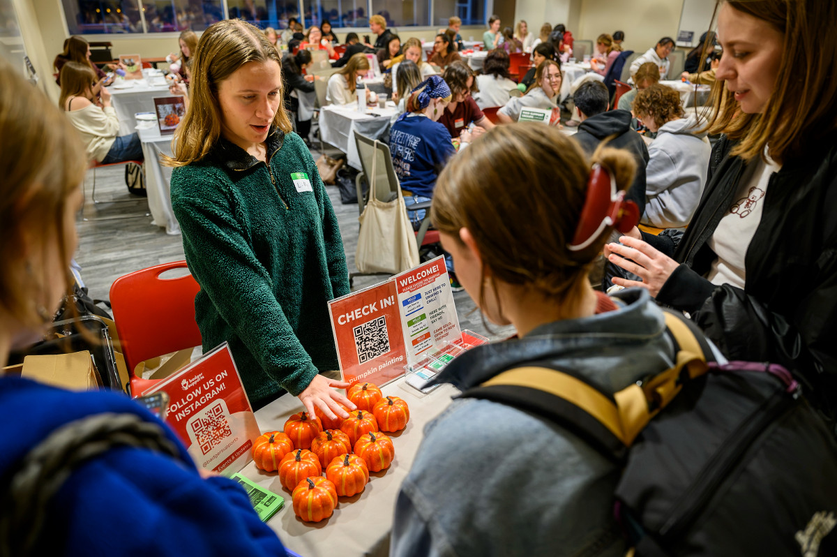 A woman stands at a check-in table speaking to a group of new-arrived students looking to get a pumpkin to paint.