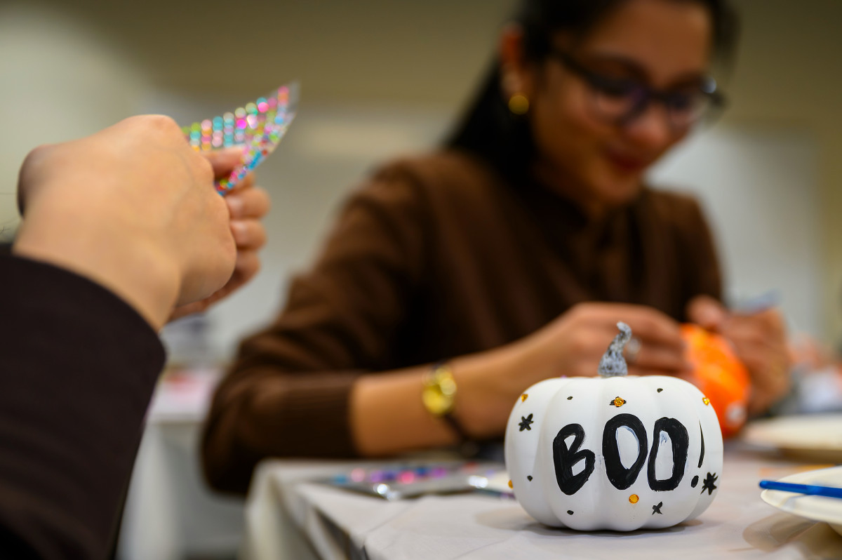 A white pumpkin painted with the word BOO rests on a table while its creator peels off gem stickers to adorn the rest of the pumpkin.
