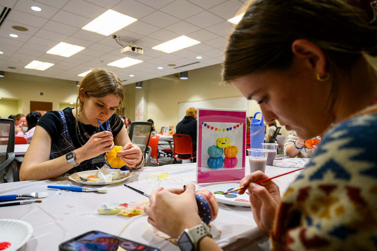Two students sit across from each other at a table where they each concentrate on painting their miniature pumpkins.