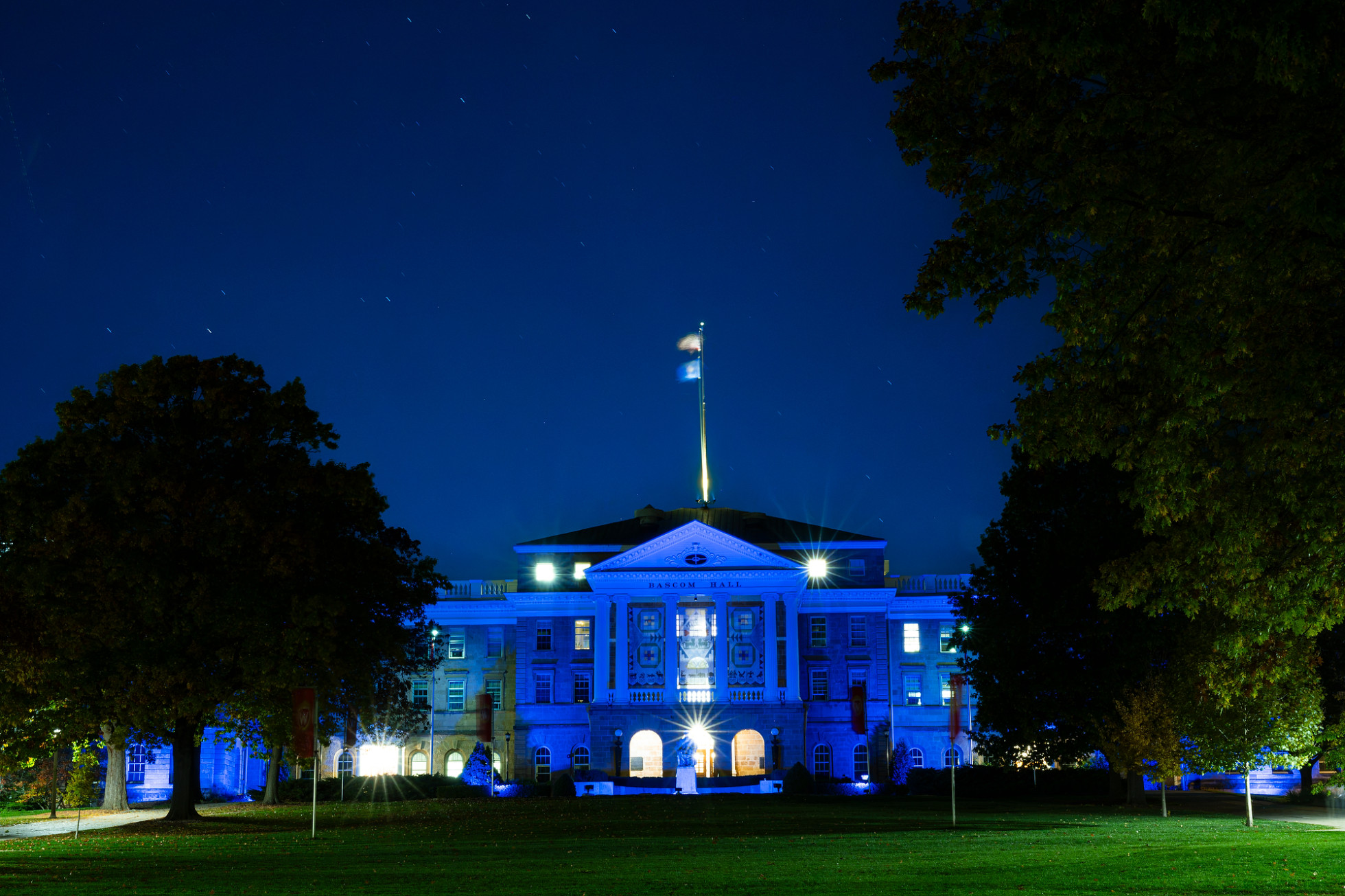 From the perspective of lower Bascom Hill, the front exterior of Bascom Hall is illuminated with blue lights at night.