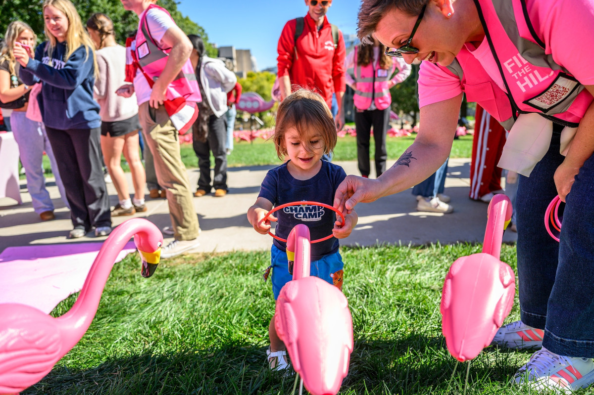 A young child tries a ring toss game over plastic pink lawn flamingos, guided by a volunteer in a pink vest.