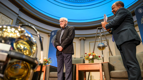 Nobel Laureate Francis Halzen smiling on a stage with blue light backdrop UW–Madison Interim Chancellor Wilcots clapping.