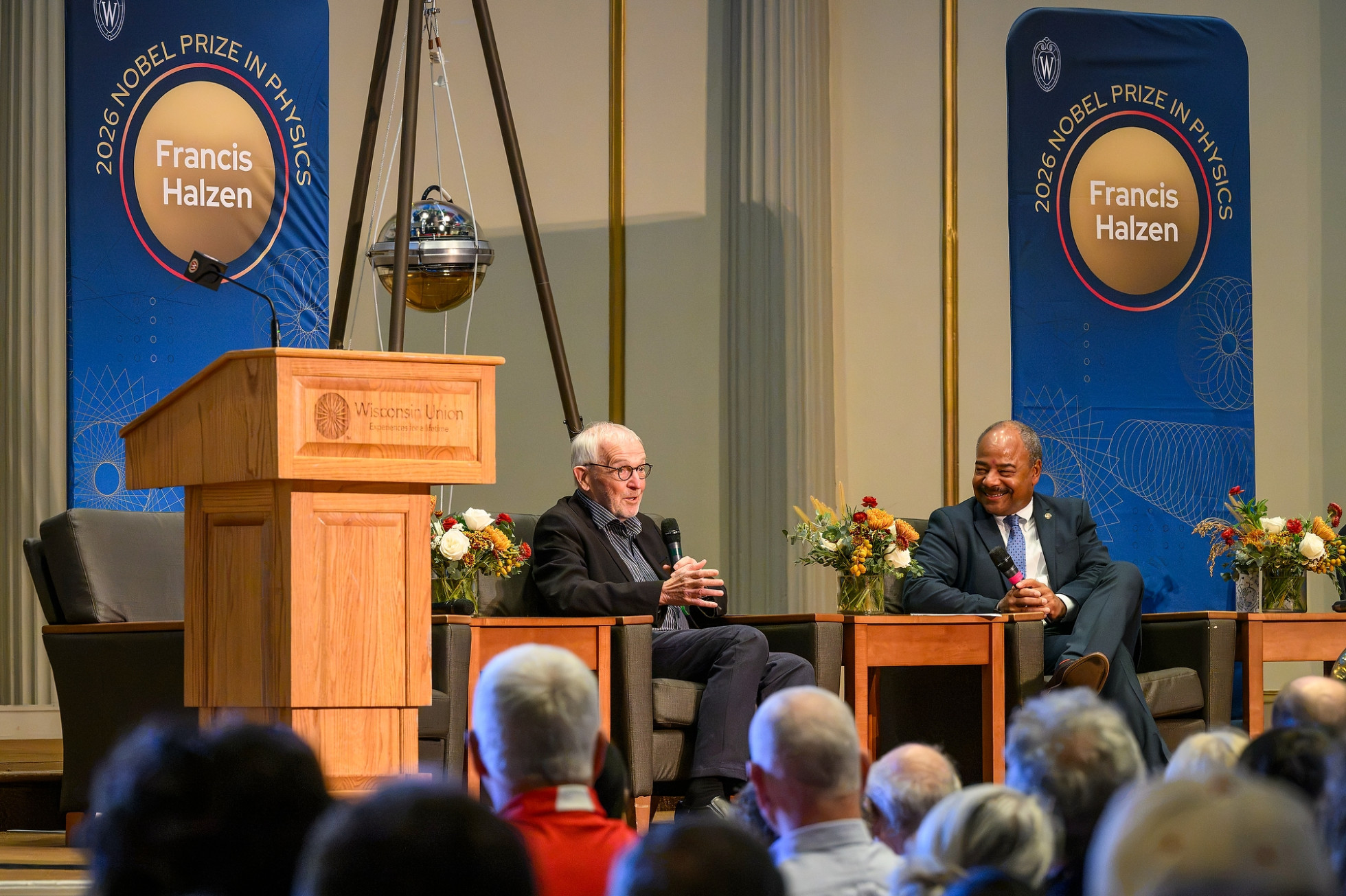 Two people sitting on a stage with microphones speaking to a crowd