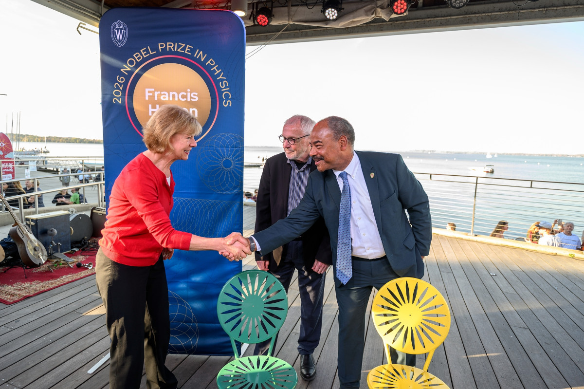 Interim Chancellor Eric Wilcots shaking hands with U.S. Senator Tammy Balwin with Nobel Laureate Francis Halzen looking on. They are pictured on an outdoor stage with Lake Mendota and a banner honoring Halzen in the background