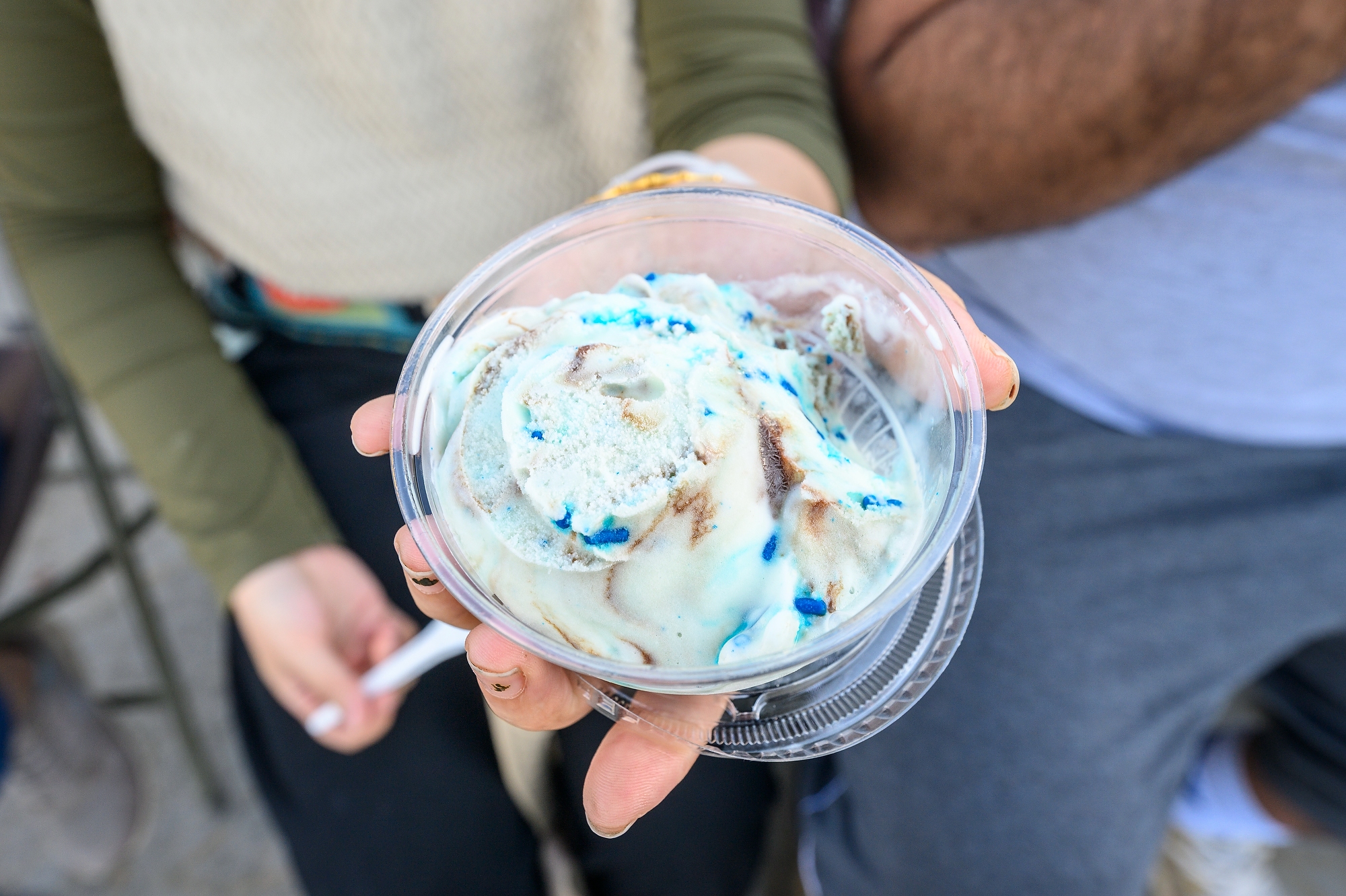A hand holding a dish of white ice cream with blue sprinkles and chocolate swirls