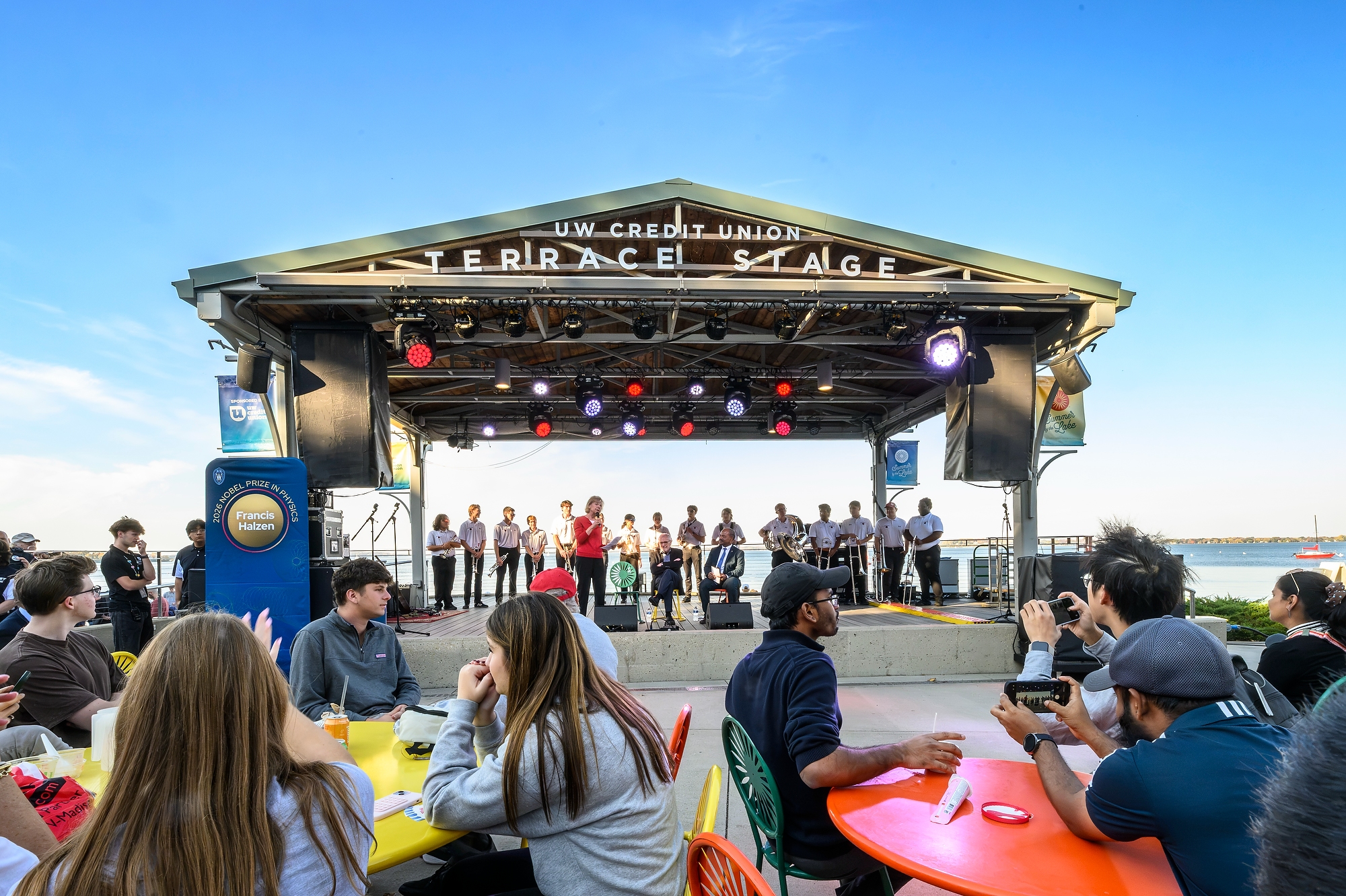 An outdoor stage with U.S. Senator Tammy Baldwin speaking. Lake Mendota pictured in the background and attendees of the Nobel Prize celebration pictured in the foreground.