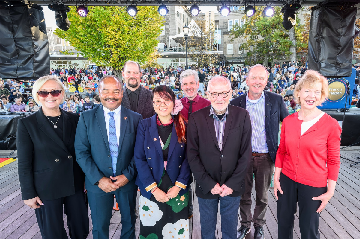 A group of people on a outdoor stage, smiling with a large crowd in the background
