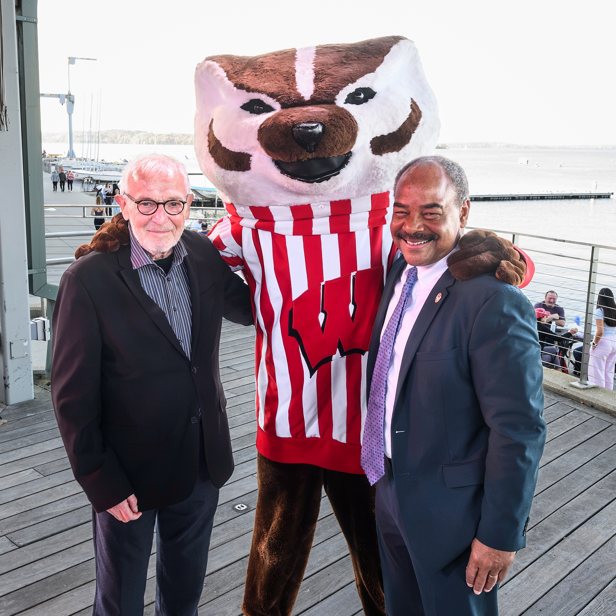 Bucky Badger mascot pictured with Nobel Laureate, Francis Halzen and Interim Chancellor Eric Wilcot on a stage with a Lake Mendota in the background.