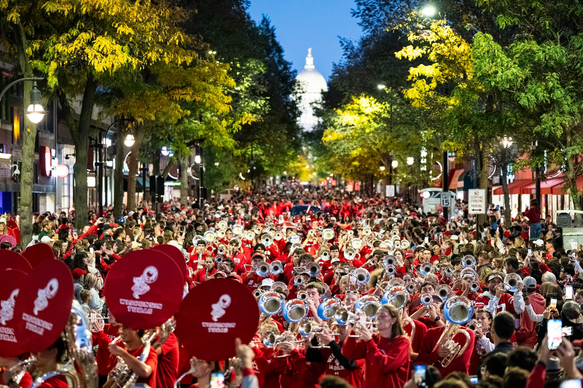 The UW Marching Band, featuring sousaphones with red covers, performs down a packed State Street during the UW Homecoming parade with the Wisconsin State Capitol in the background.