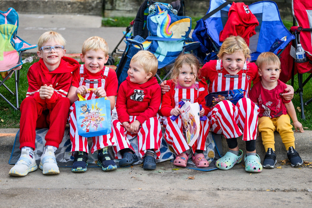 Six children wearing matching red outfits and red-and-white striped bib overalls sit together along the curb waiting for the UW Homecoming parade.