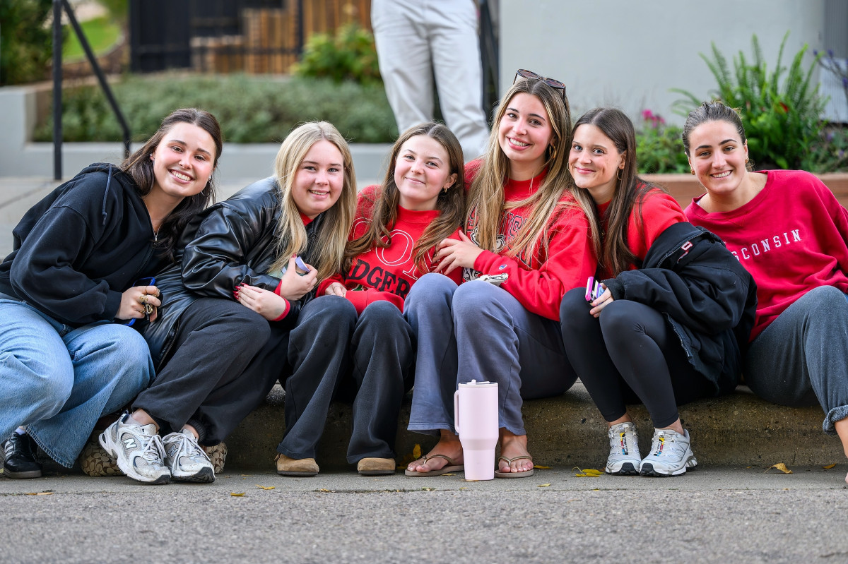 A group of six young women dressed in Wisconsin sweatshirts sit side-by-side along the sidewalk curb, smiling for the camera.