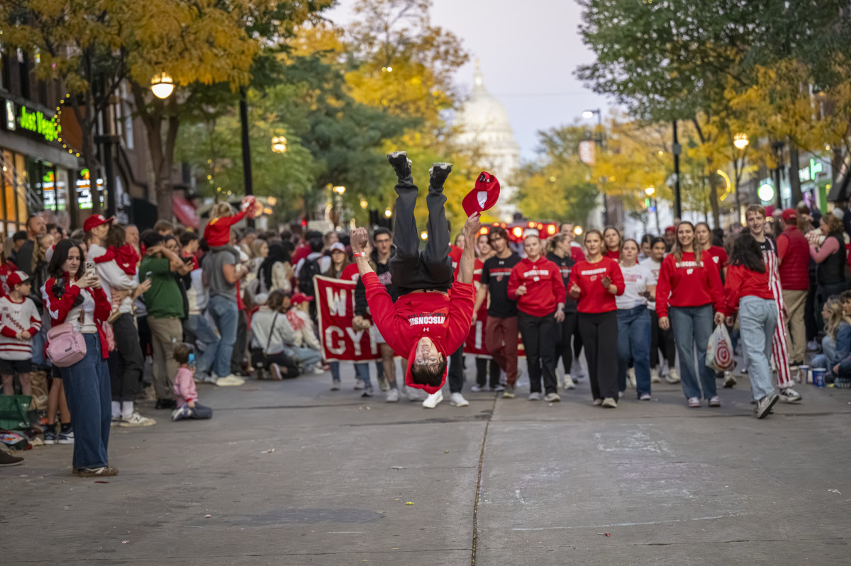 A person in a red Wisconsin Badgers sweatshirt performs a backflip on a crowded street during a lively parade. Spectators watch and cheer, many dressed in red attire. The Wisconsin State Capitol building is visible in the background.