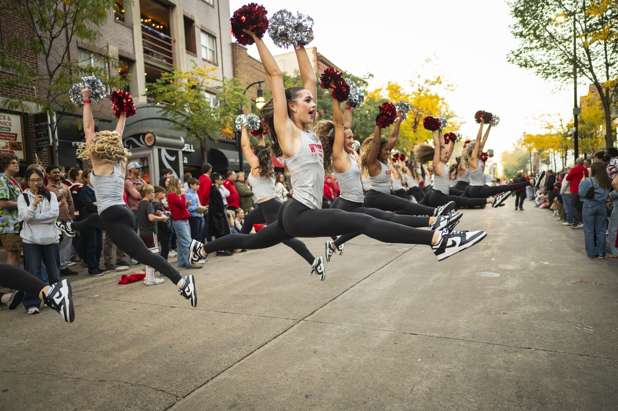 A group of dancers perform energetic jumps, each holding red and silver pom-poms, during a street parade lined with onlookers.
