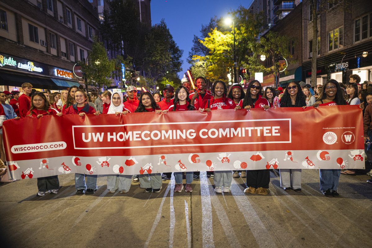 Students stand in a line one State Street holding a red banner that reads UW Homecoming Committee.
