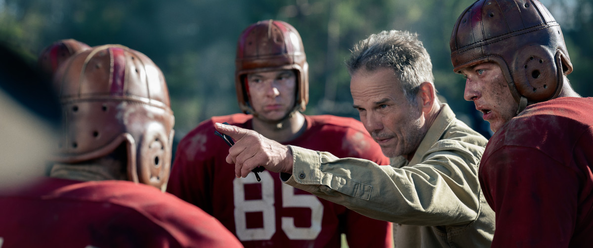 A man points to a location in the distance while talking to a group of actors wearing replica 1940's Wisconsin Badgers football uniforms.