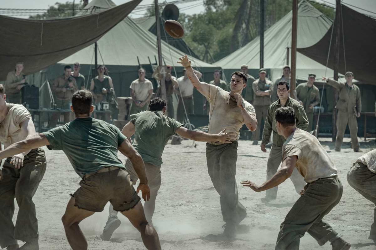 Soldiers in vintage uniforms play football on a dusty field surrounded by military tents, with other soldiers watching, suggesting a break in a wartime or military camp setting.
