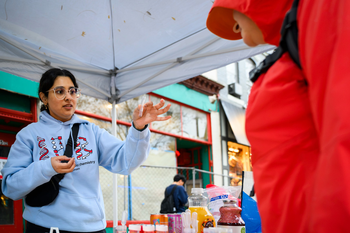 Standing under a tent on State Street, a woman holds a small glass vial in her hand and shows its contents to an observer.