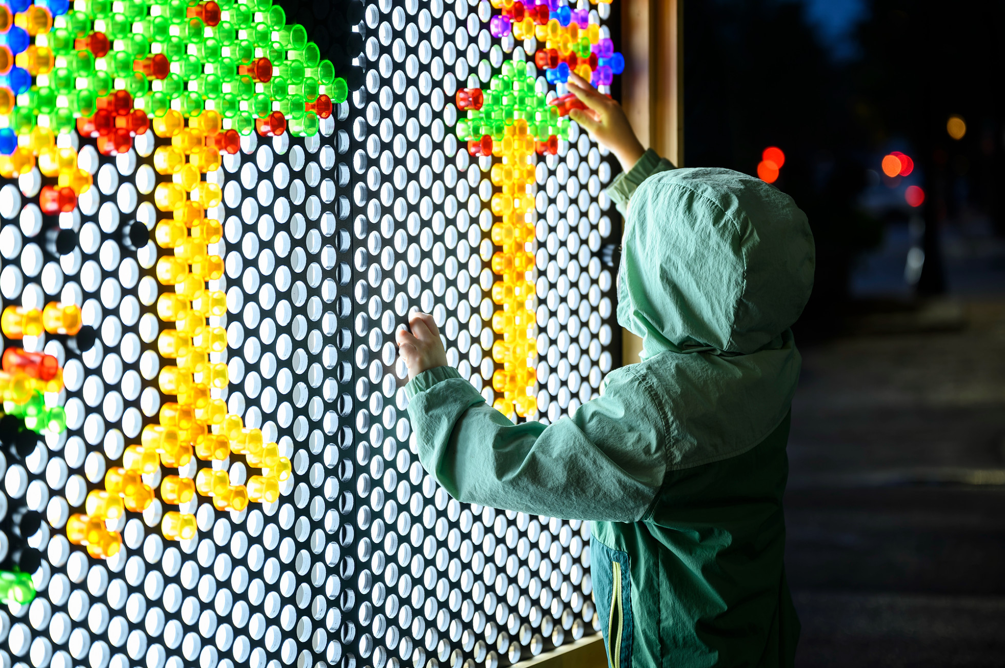 A young child wears a green rain jacket while adding brightly colored pegs into a huge light-up peg board.