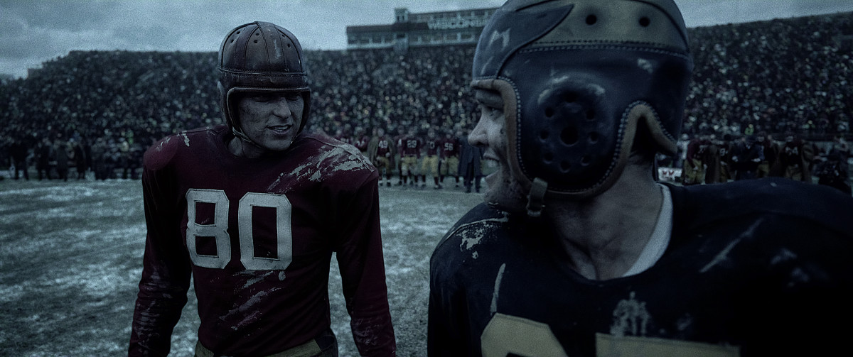 An actor wears a replica of a 1940's Wisconsin football uniform that displays the number 80 while talking to another actor wearing a replica Purdue jersey.